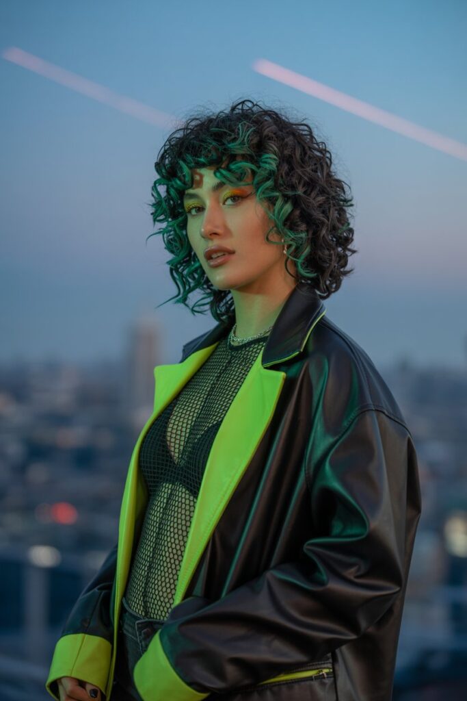 A bold curly-haired woman with shoulder-length black curls and a vivid emerald green skunk stripe framing her face